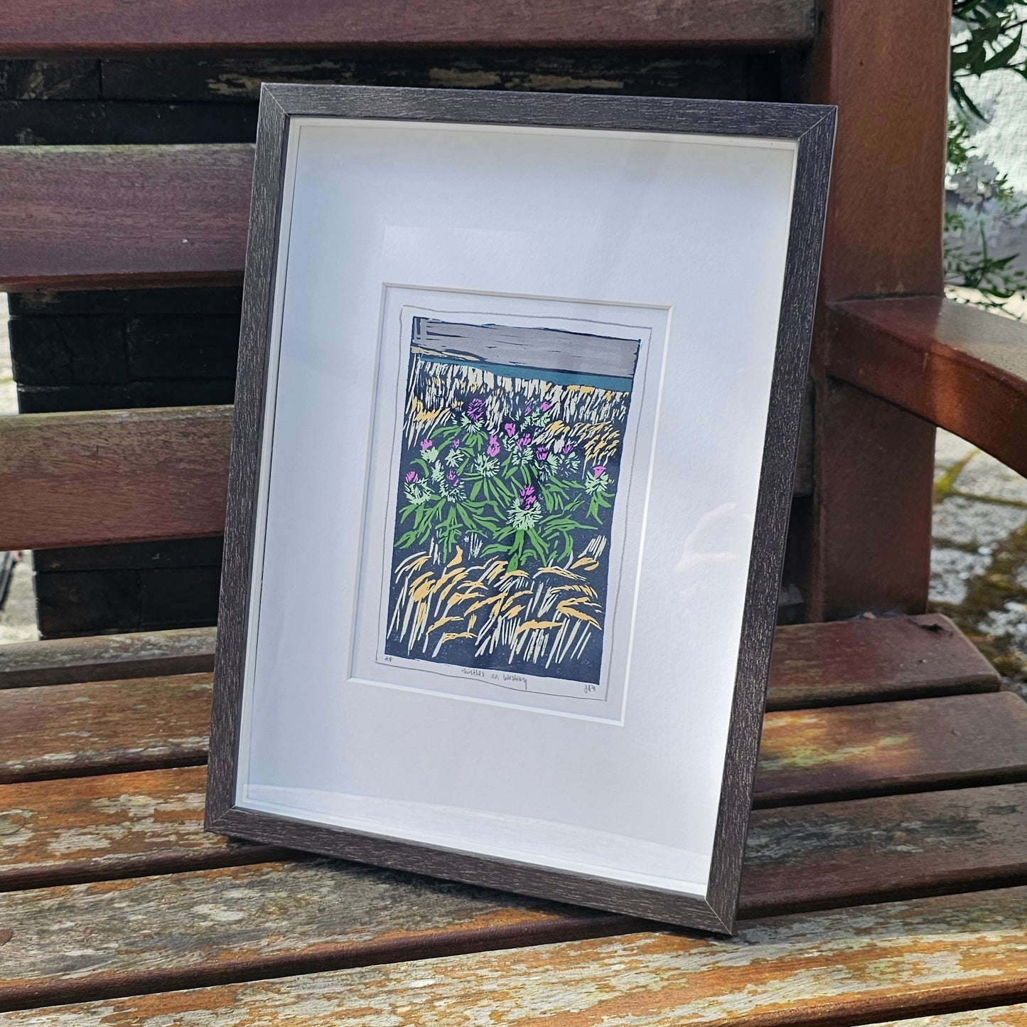 A colourful framed lino print of Scottish Westray thistles by BeeBirds, with a black/brown frame, set against a background of a wooden slatted bench.