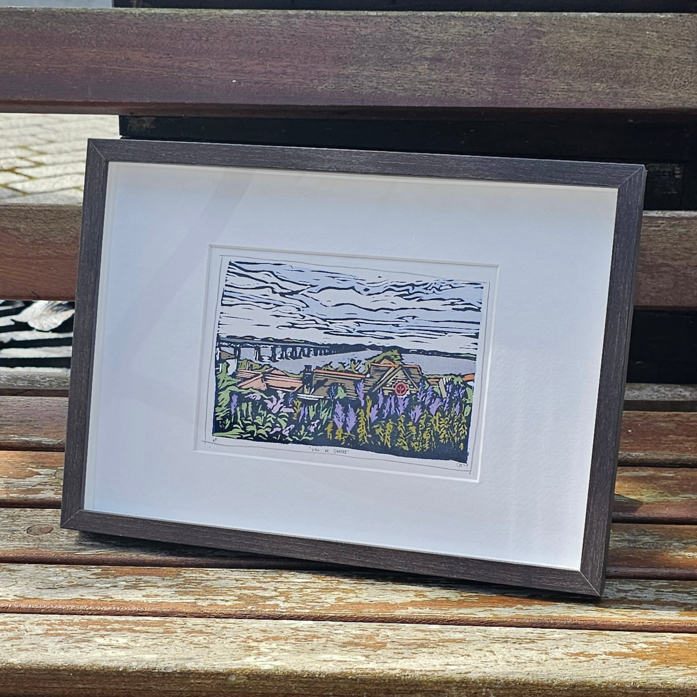 A colorful lino print of 'View To Dundee' framed in a black frame, placed on a wooden surface.