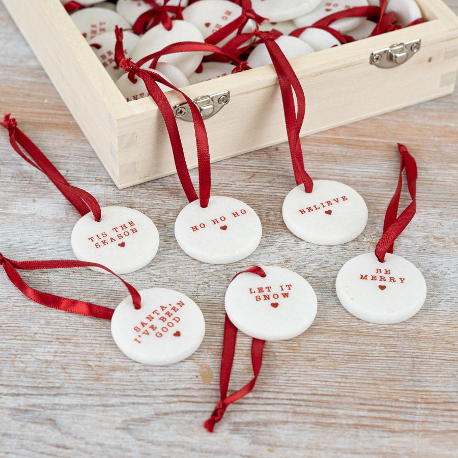 A set of six small round white ceramic Christmas pebble decorations with red text and a red ribbon.