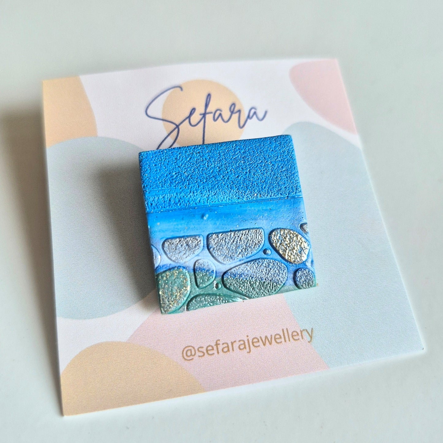 Blue sand textured square brooch on a colourful card with 'Sefara' branding