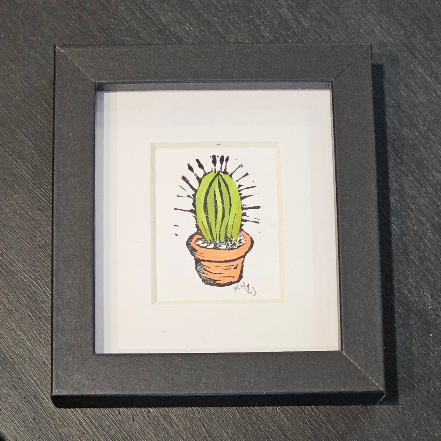 A colourful lino print of a cactus in a small black frame, placed on a black surface