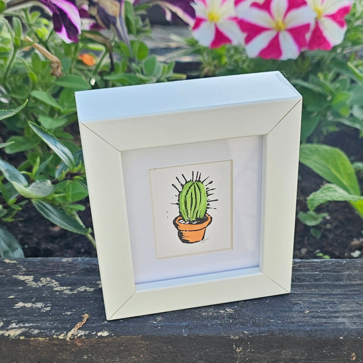A colourful lino print of a cactus in a small white frame, placed on a wooden surface with plants in the background.