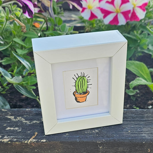 A colourful lino print of a cactus in a small white frame, placed on a wooden surface with plants in the background.