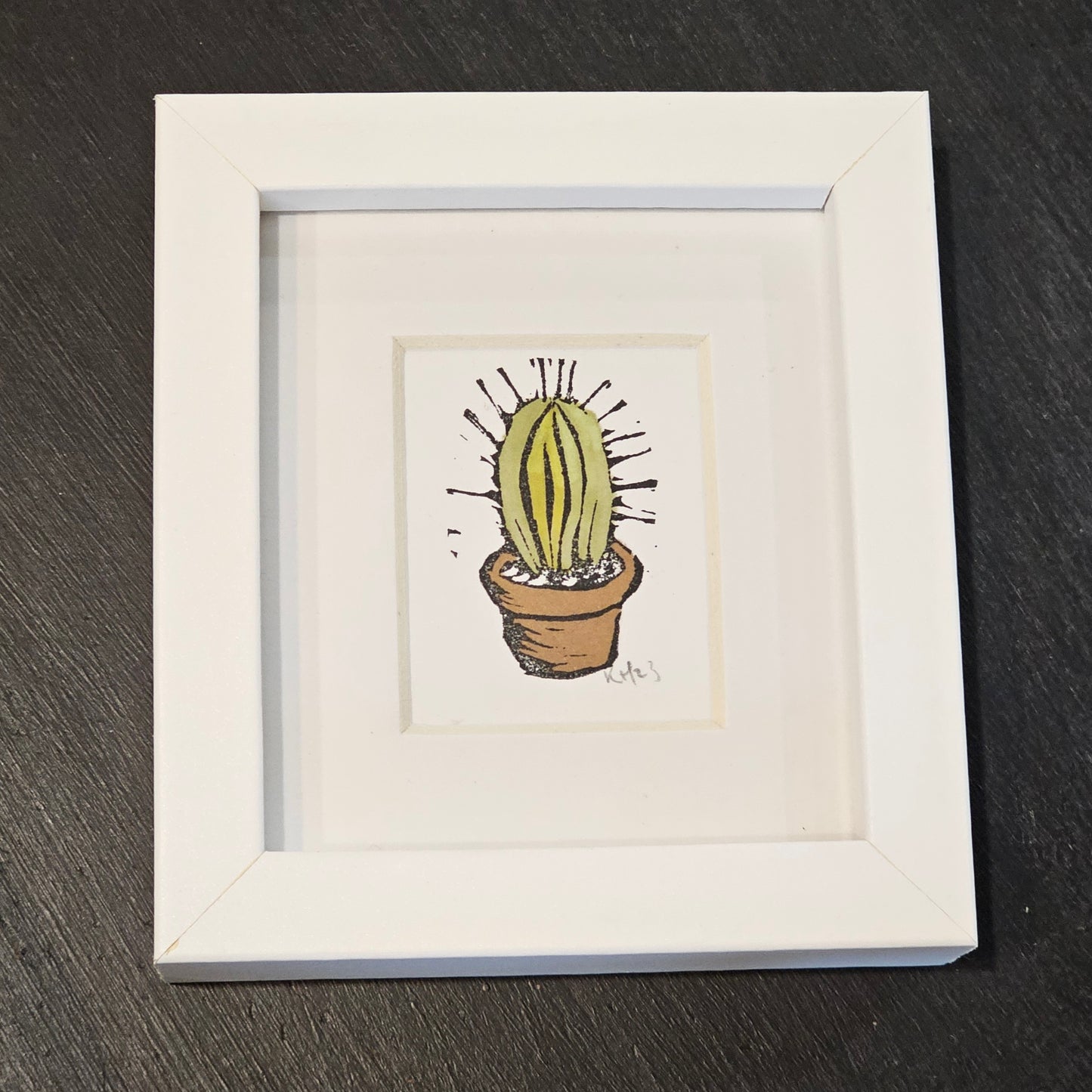 A colourful lino print of a cactus in a small white frame, placed on a black surface