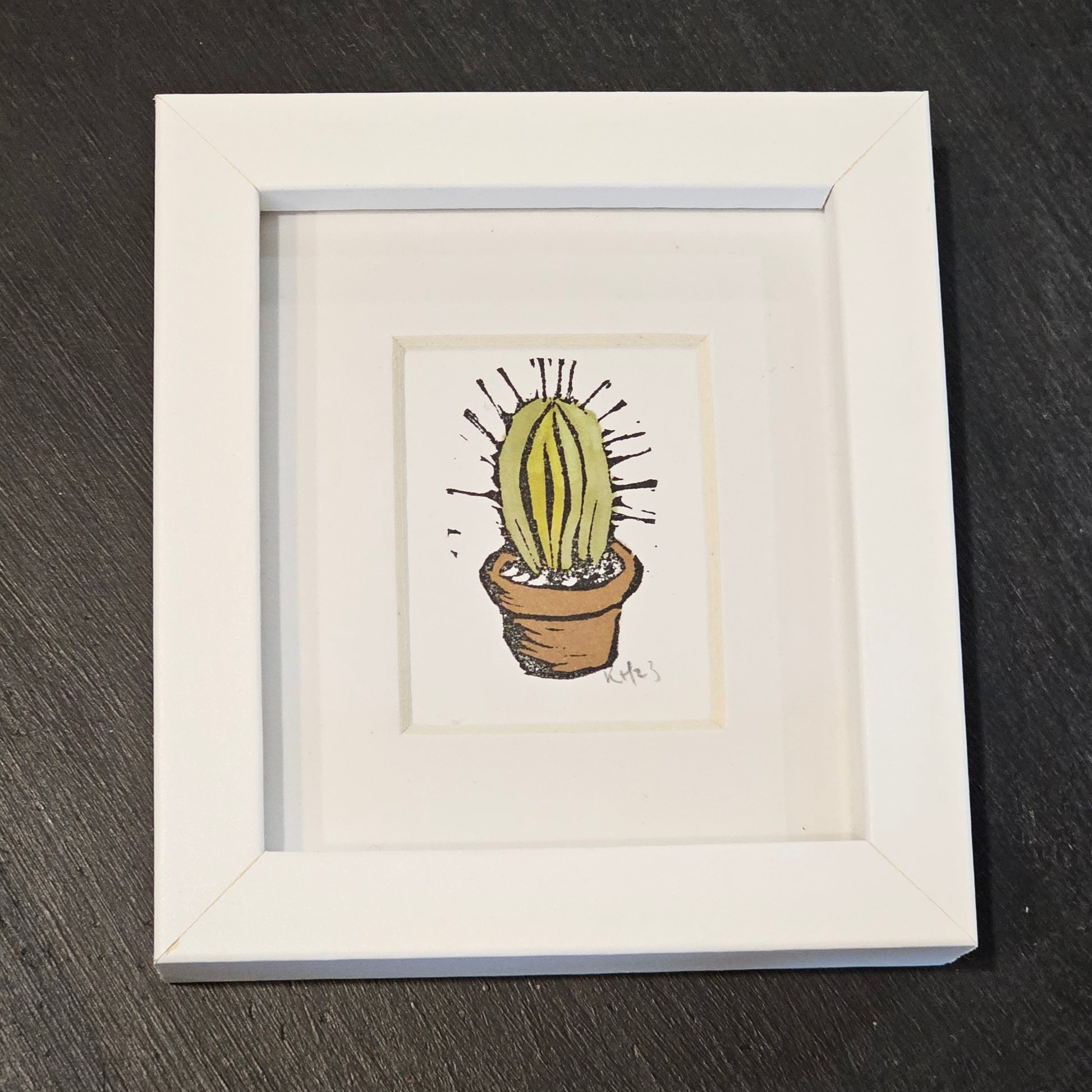 A colourful lino print of a cactus in a small white frame, placed on a black surface