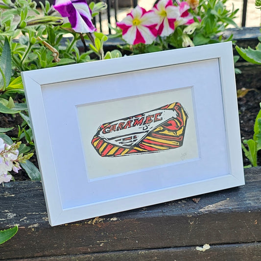 Framed Colourful Caramel Wafer Lino Print by Bee Birds on wooden surface with flowers in background