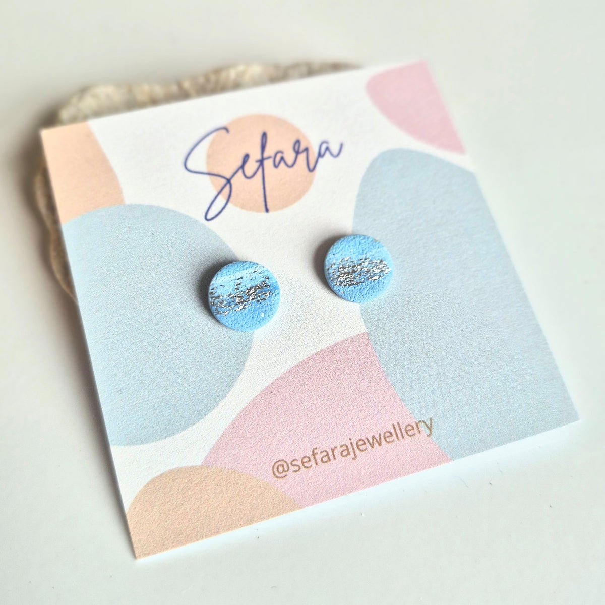 Blue round stud earrings with silver accents on a colourful card with 'Sefara' branding