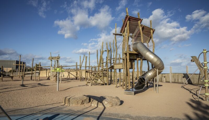 Children's playground with wooden structures and a slide on a sandy area under a blue sky.