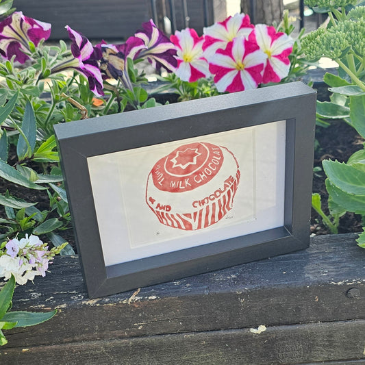 A colorful lino print of a teacake in a black frame, displayed on a wooden surface with flowers in the background.