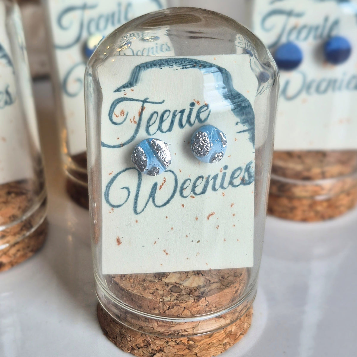 Sefara Jewellery Teenie Weenie Coastal Stud Earrings featuring miniature seascape designs with gold or silver leaf detailing, presented in glass cloche domes with cork stoppers.