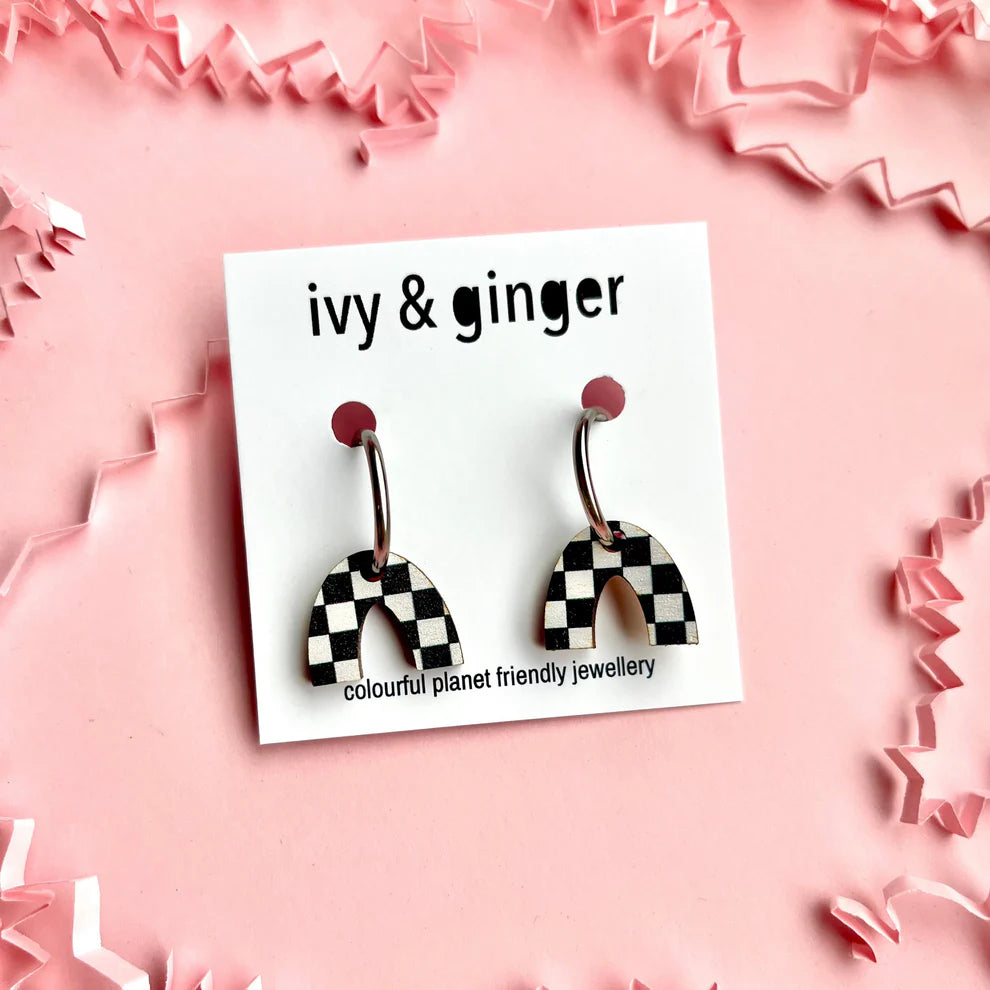 A pair of checkerboard print arch drop stud earrings displayed on a pink background, with the brand name 'Ivy & Ginger' on the packaging.