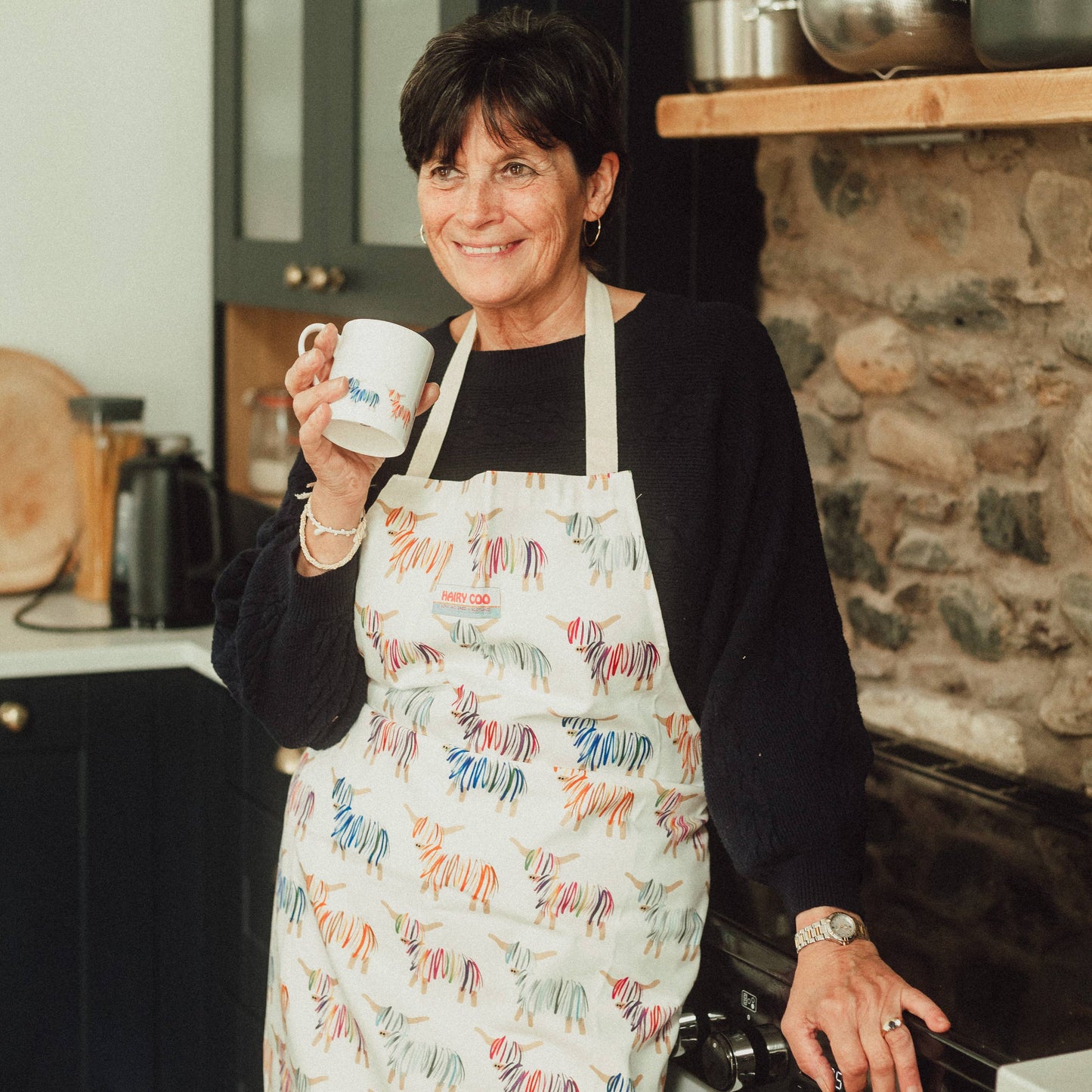 Woman in a kitchen wearing Hairy Coo 100% cotton apron with bright Highland Cow print and drinking from a Hairy Coo mug