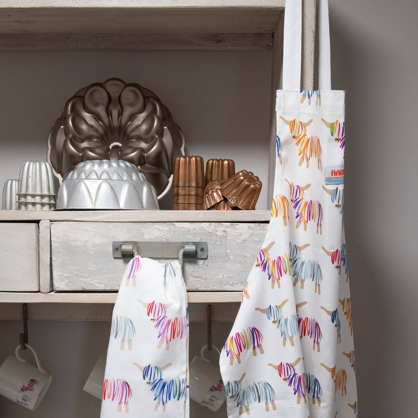 Hairy Coo 100% cotton apron with bright Highland Cow print hangng next to a Hairy Coo teatowel