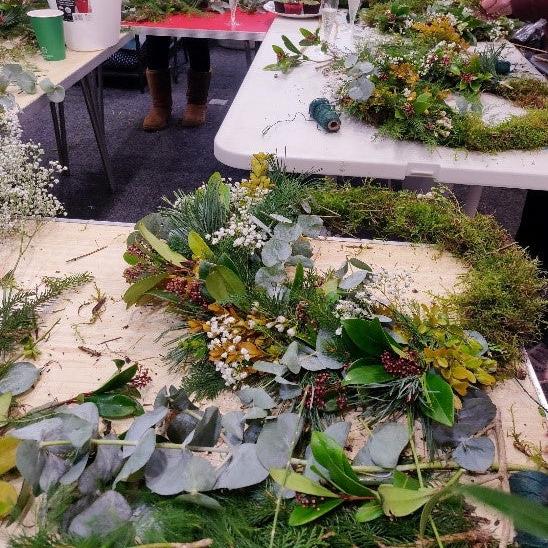 A close-up of fresh, green foliage and natural decorations onto a willow and moss wreath base during a workshop.