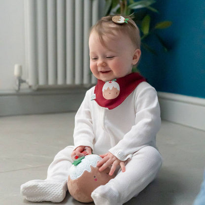 Baby sitting on the floor wearing a white outfit and red L'il Pudding Christmas baby bandana bib