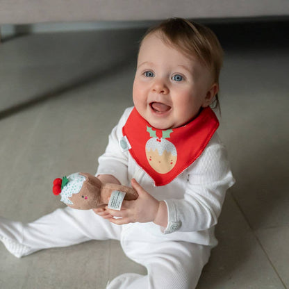Baby wearing a red bib with reindeer design, holding a 1st Christmas pudding plush baby rattle