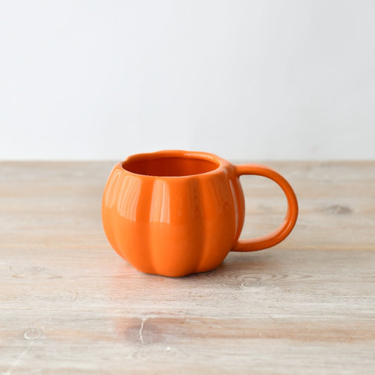 A glossy orange ceramic pumpkin-shaped mug with a handle, placed on a wooden surface against a white background.