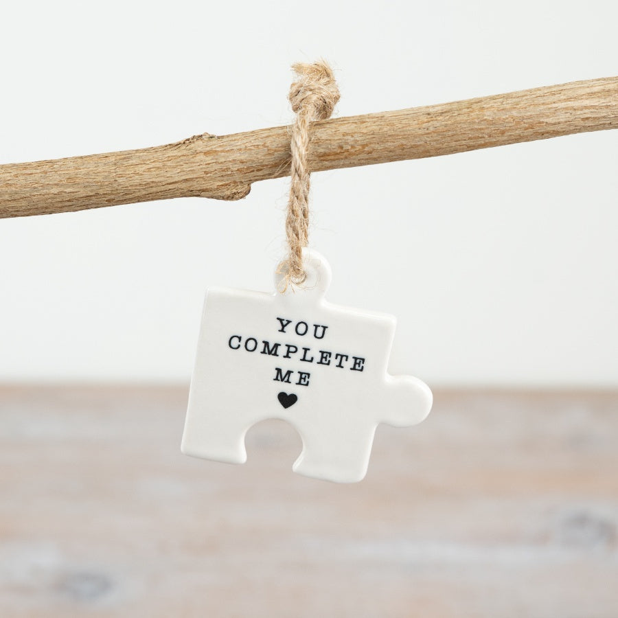 A small hanging ceramic jigsaw puzzle piece with the words "you complete me" and a heart symbol.