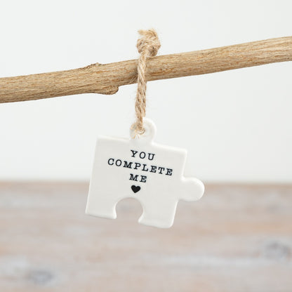 A small hanging ceramic jigsaw puzzle piece with the words "you complete me" and a heart symbol.