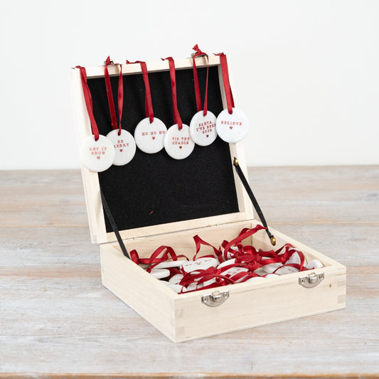 A set of six small round white ceramic Christmas pebble decorations with red text and a red ribbon. 