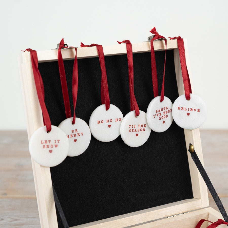 A set of six small round white ceramic Christmas pebble decorations with red text and a red ribbon. 