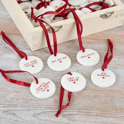 A set of six small round white ceramic Christmas pebble decorations with red text and a red ribbon. 