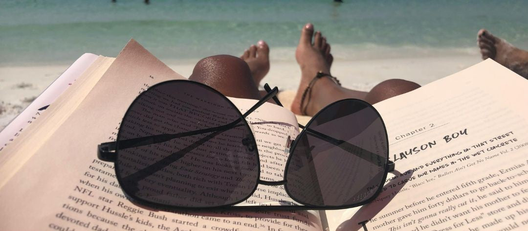 Black round-frame sunglasses resting on an open book at the beach, with a person's bare feet visible in the sand near turquoise water.