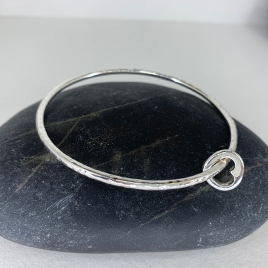 A single thick silver bangle with a small cutout heart charm, displayed on a stone surface.