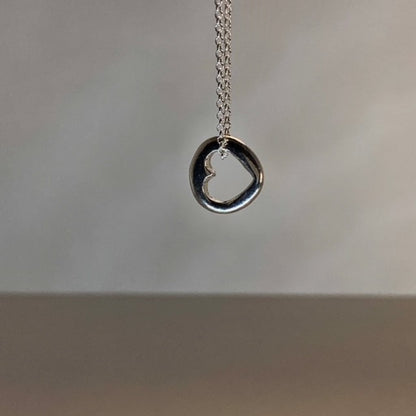 Close-up of a small, round solid sterling silver pendant with a central cut-out heart, approximately 1.2cm in diameter, suspended on an 18-inch silver chain.