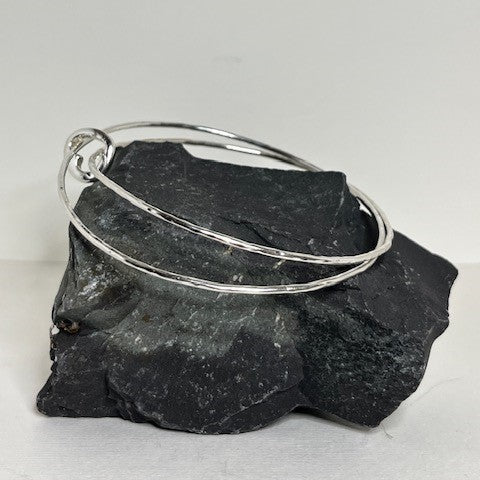 A double silver bangle with a small cutout heart charm, displayed on a stone surface.