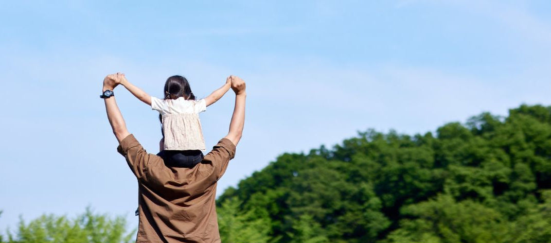 Father with child on shoulders against a blue sky with green trees
