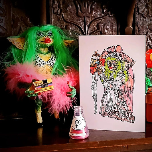 A printed card with an illustration of a Greta the gremlin bride next to a Greta toy with green hair and pink feathers.