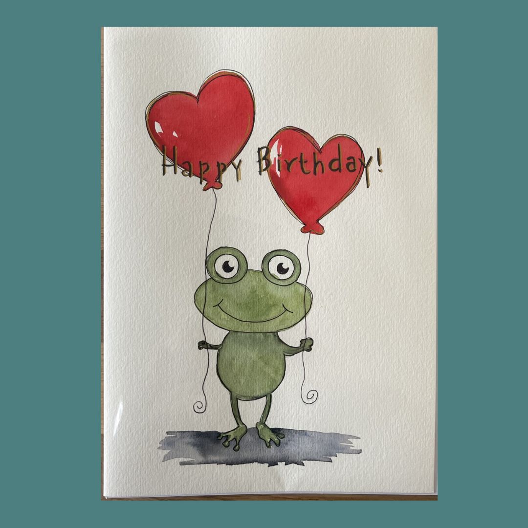 A greeting card featuring a watercolor-style green frog holding two red heart-shaped balloons with the words 'Happy Birthday!' written across the top