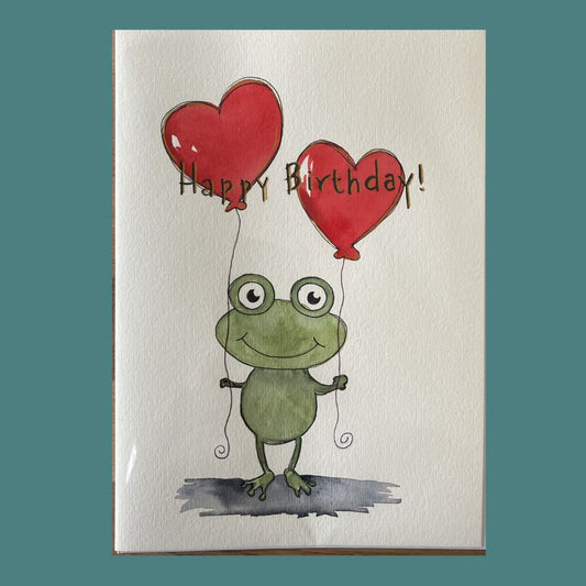 A greeting card featuring a watercolor-style green frog holding two red heart-shaped balloons with the words 'Happy Birthday!' written across the top