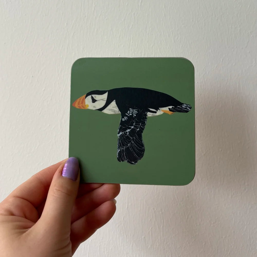 Hand holding a square coaster with a puffin design against a plain background