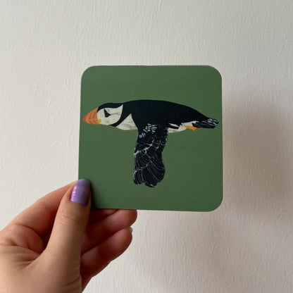 Hand holding a square coaster with a puffin design against a plain background