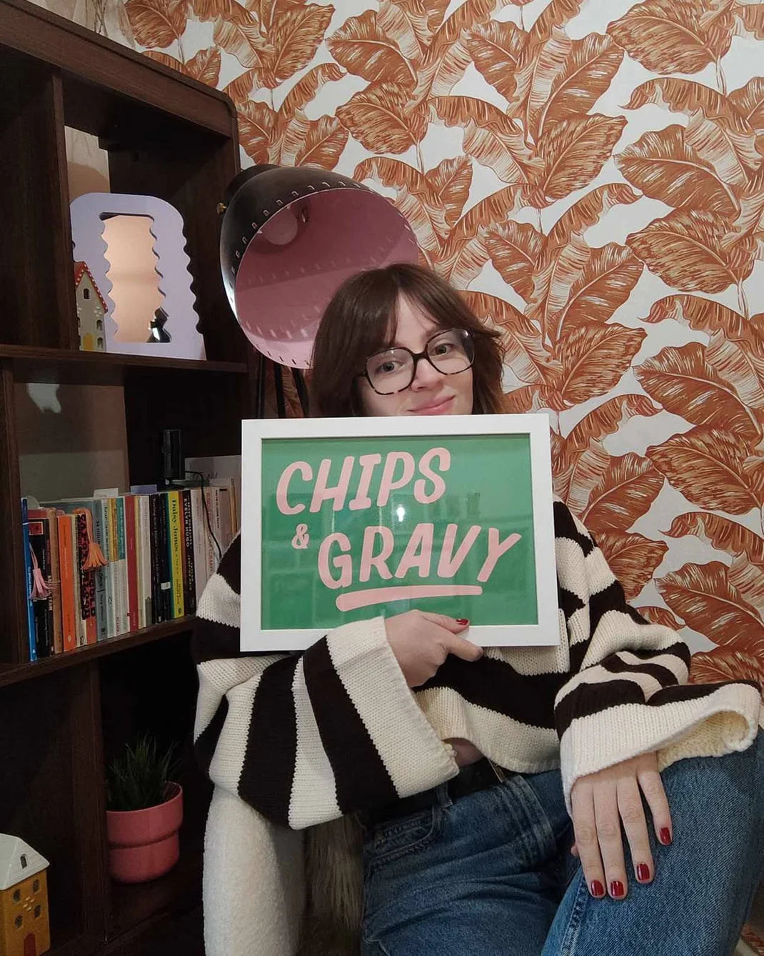 Laure Beth holding a framed poster saying 'Chips & Gravy' in a room with bookshelf and decorative wall.