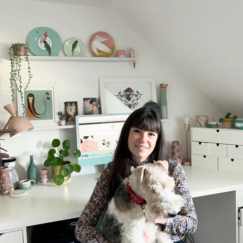 Mica sitting at a desk with a dog, surrounded by decorative items and plants.