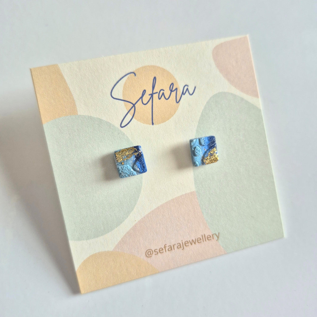 Blue square stud earrings with gold detail on a colourful card with Sefara branding