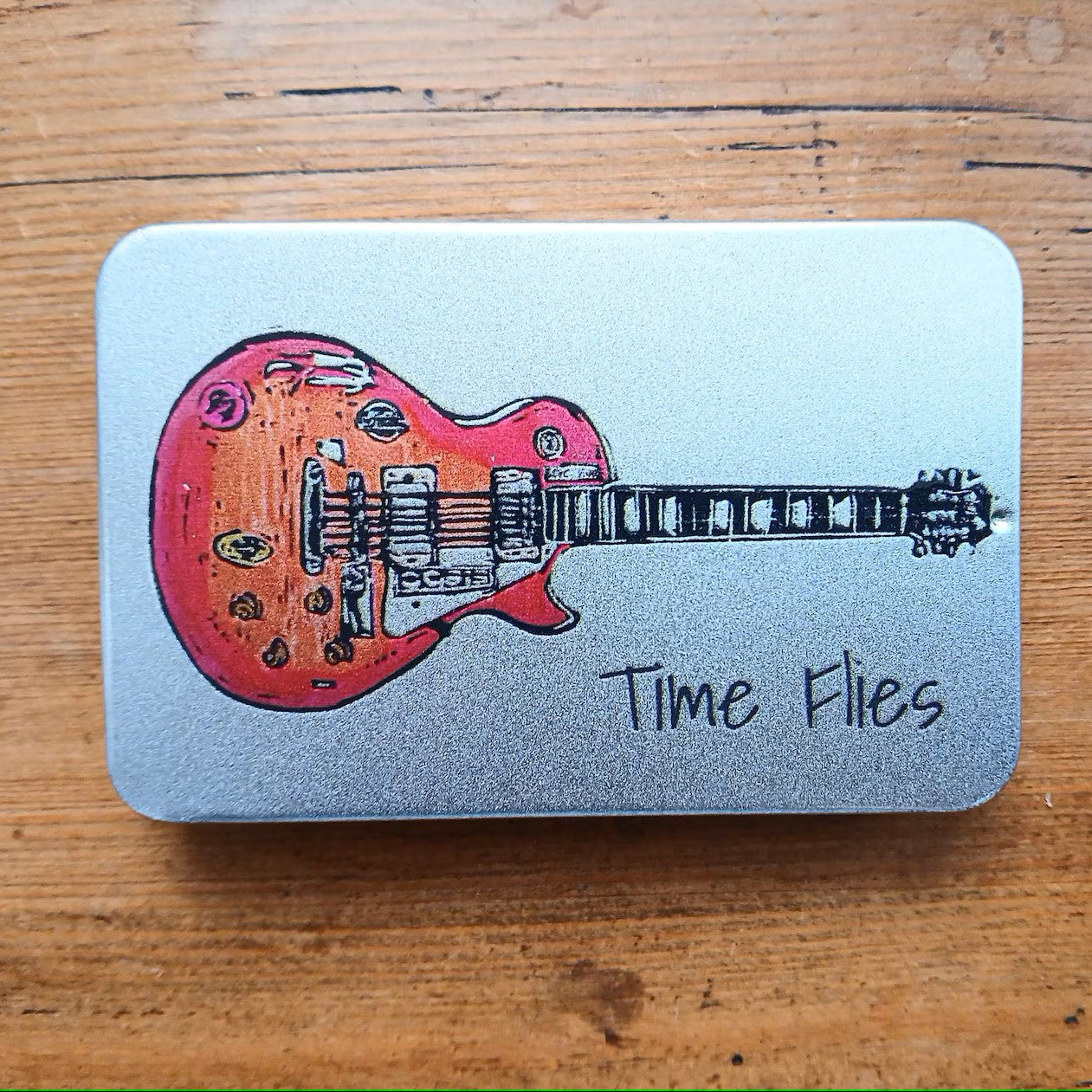 Silver rectangular tin with sliding lid featuring a guitar image with the phrase 'Time Flies' written on it.