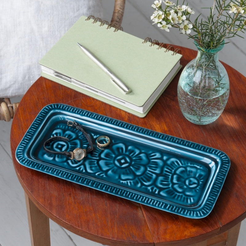 Teal blue enamel jewellery tray on a wooden table with a notebook and vase in the background