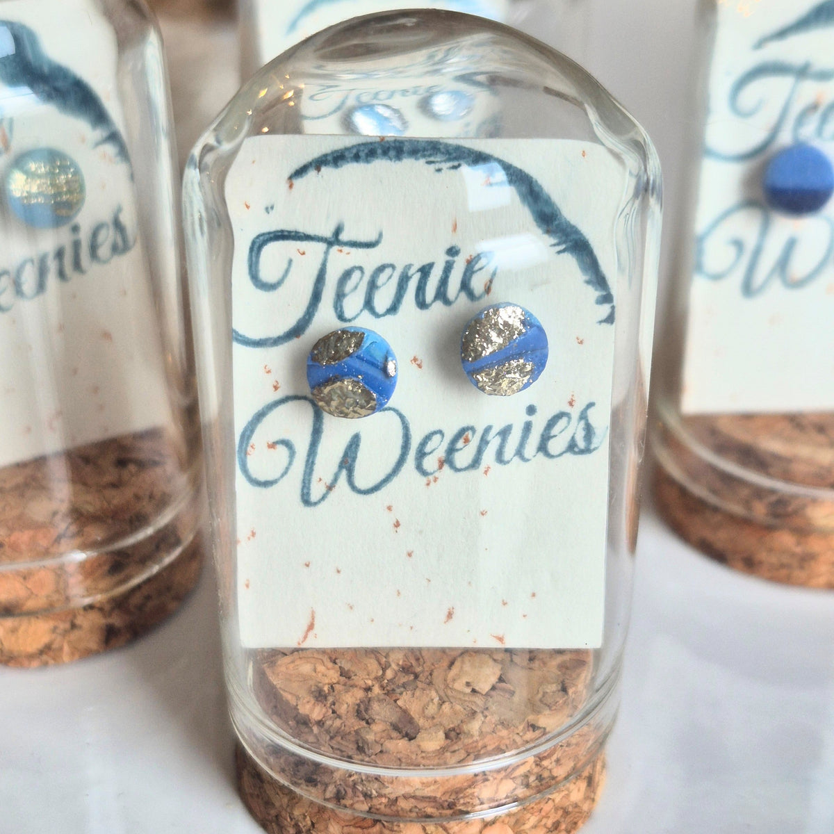 Sefara Jewellery Teenie Weenie Coastal Stud Earrings featuring miniature seascape designs with gold or silver leaf detailing, presented in glass cloche domes with cork stoppers.