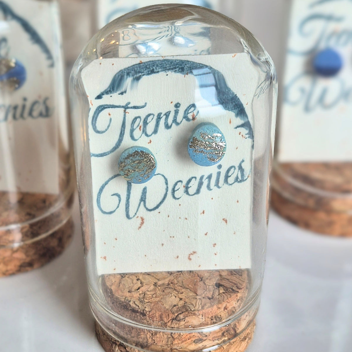 Sefara Jewellery Teenie Weenie Coastal Stud Earrings featuring miniature seascape designs with gold or silver leaf detailing, presented in glass cloche domes with cork stoppers.