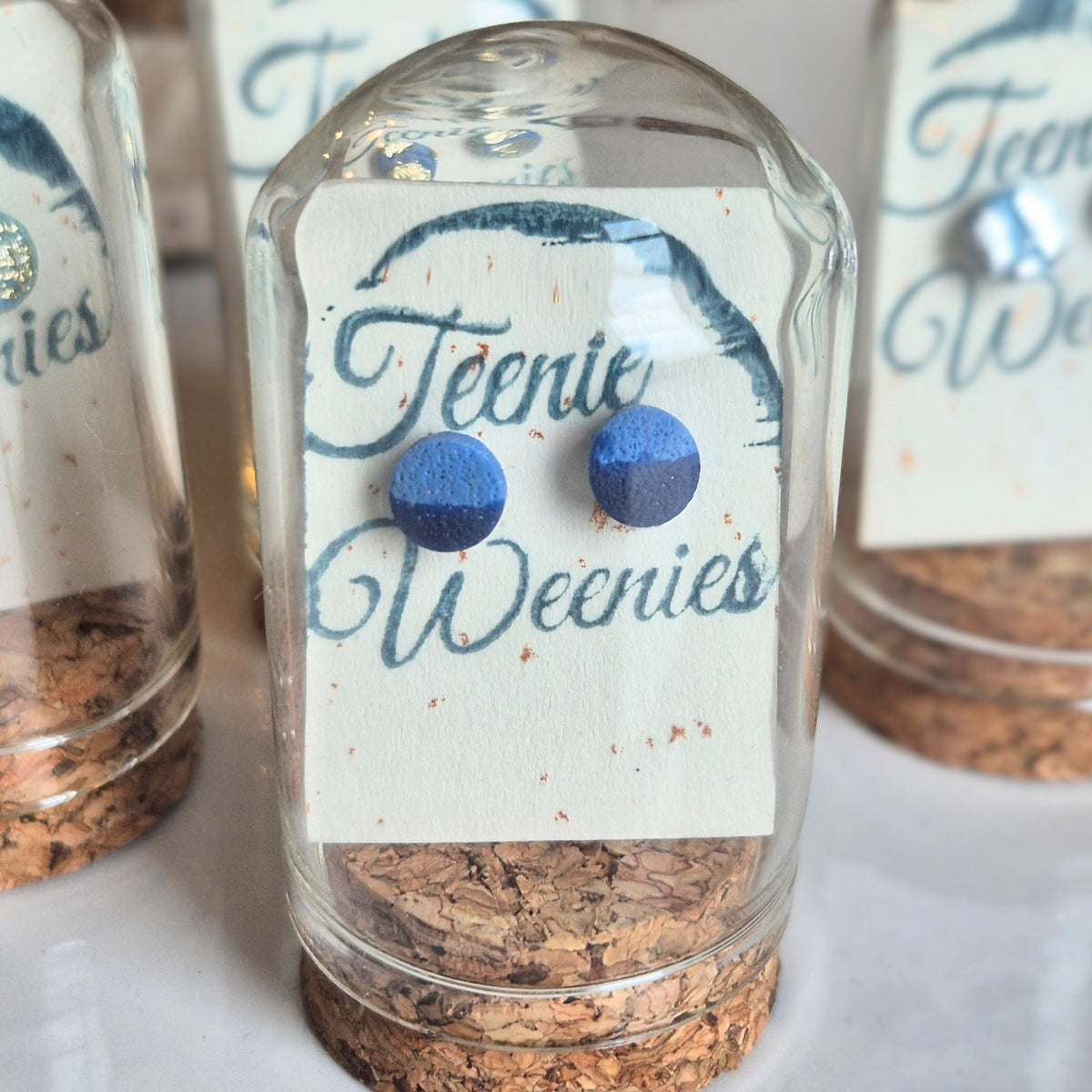 Sefara Jewellery Teenie Weenie Coastal Stud Earrings featuring miniature seascape designs with gold or silver leaf detailing, presented in glass cloche domes with cork stoppers.