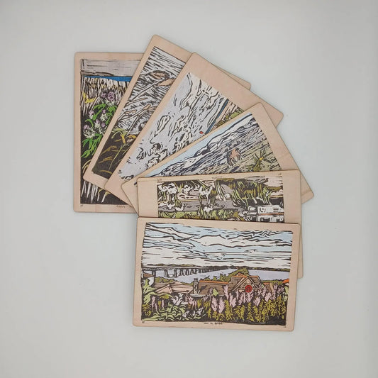 A collection of postcards with wooden postcard bases, each featuring a unique black and white lino cut image of a cycling-related scene or landscape.