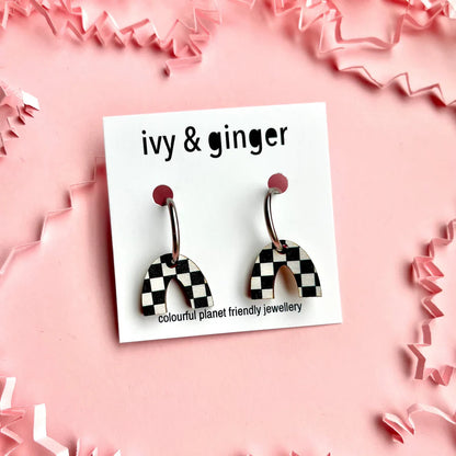 A pair of checkerboard print arch drop stud earrings displayed on a pink background, with the brand name 'Ivy & Ginger' on the packaging.